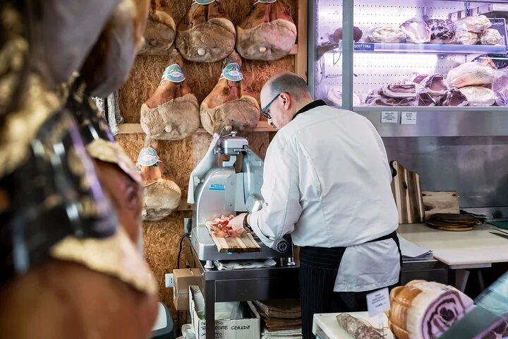 A man in a white jacket slicing cured meat on a slicer in a deli, with whole hams hanging on the wall behind
