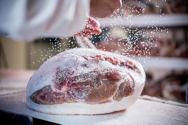 Eine Hand streut grobes Meersalz über eine frische Schweinekeule auf einem weißen Tisch