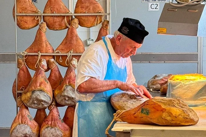 A ham maker in a black cap and blue apron trimming a cured ham on a table, with hams hanging on racks behind him